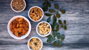 Aerial view of assorted nuts and seeds in white bowls with green leaves on a rustic wooden table.